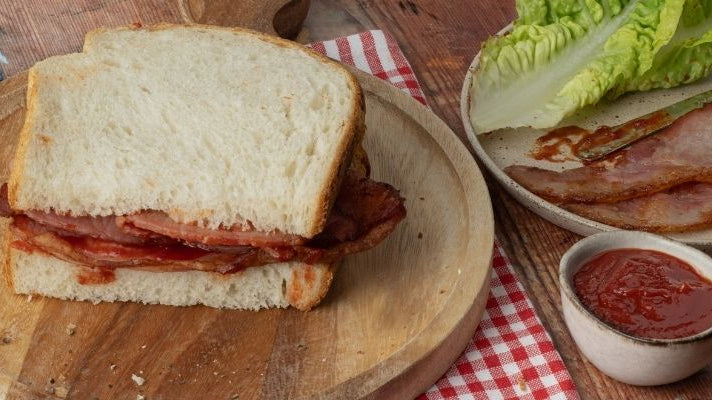 A bacon sandwich on white bread with a small pot of Tiptree Tomato Ketchup and lettuce off to one side.