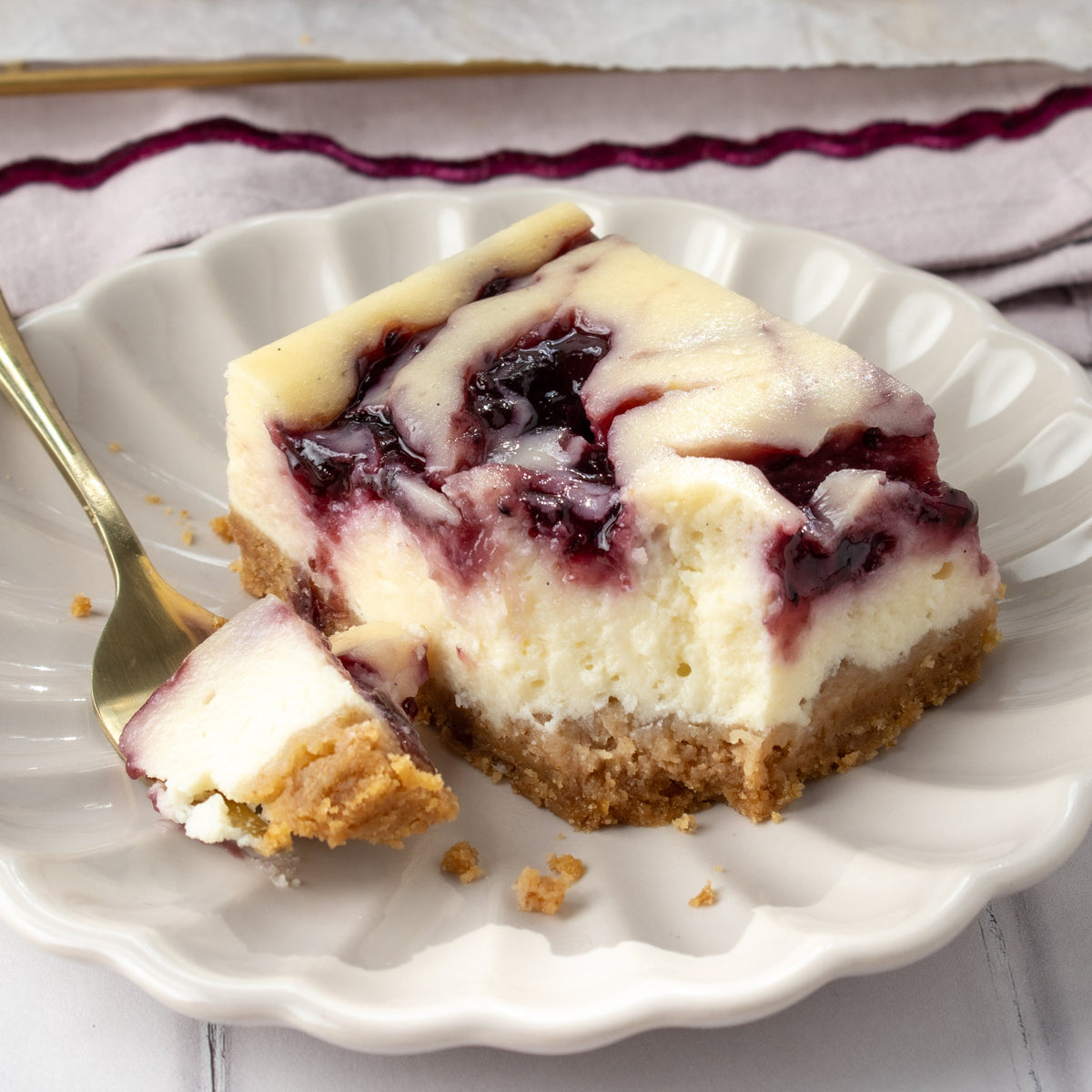 A close-up of a slice of Black Currant cheesecake on a white scalloped plate with a gold fork, showing the creamy white filling, dark Black Currant topping, and biscuit base.