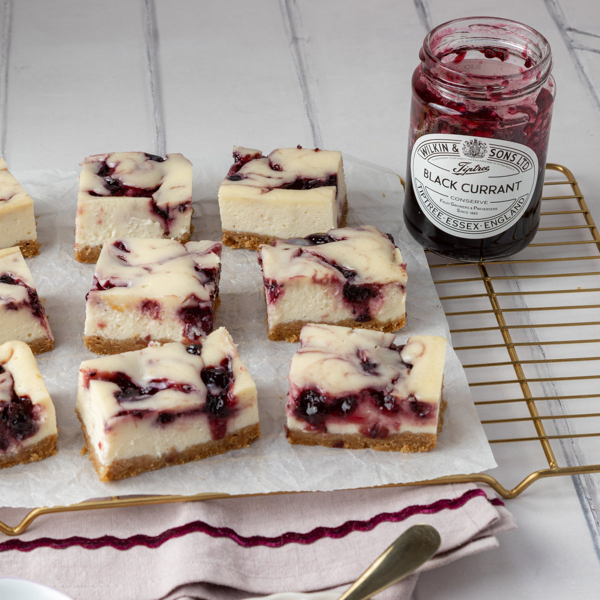 Several square cheesecake bars with a buttery biscuit base and dark Black Currant swirls are arranged on parchment paper on a gold wire cooling rack, next to a jar of Wilkin & Sons Tiptree Black Currant Jam.