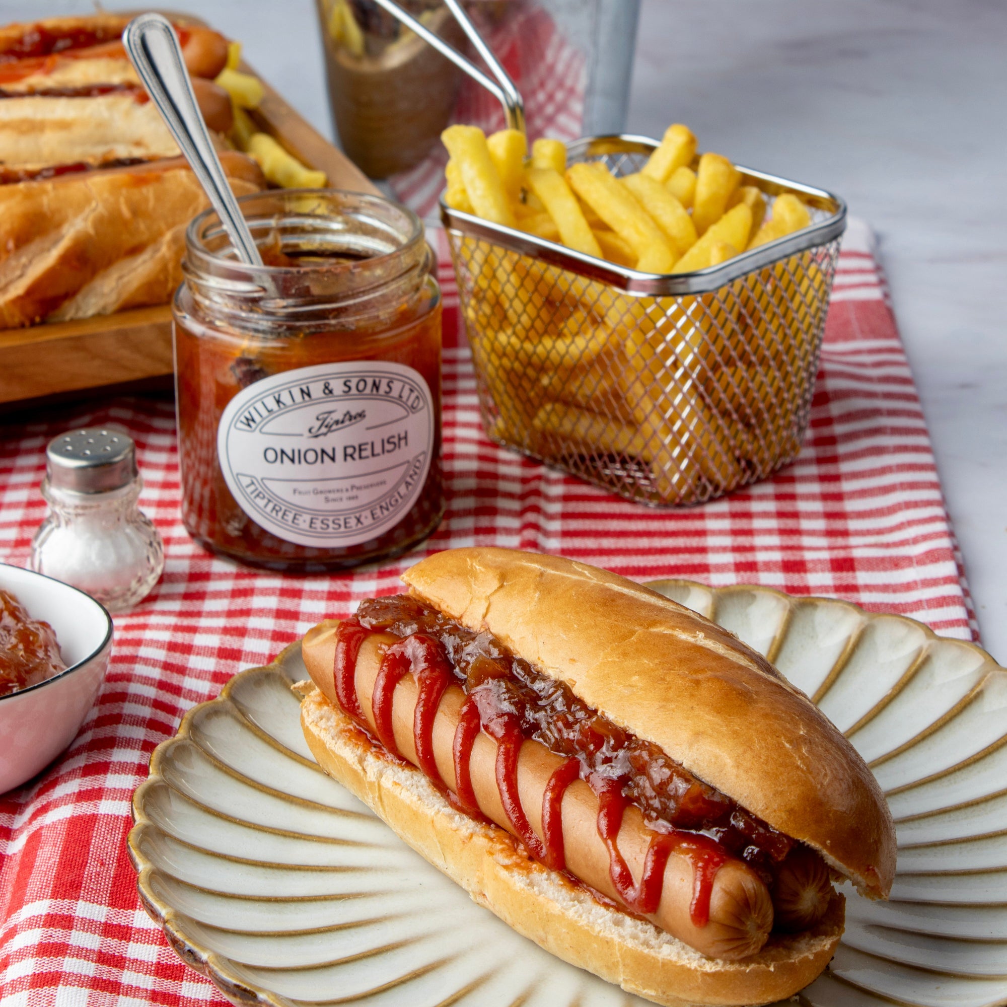 Hot dog with relish on a bun, accompanied by fries and onion relish jar on a checkered tablecloth.