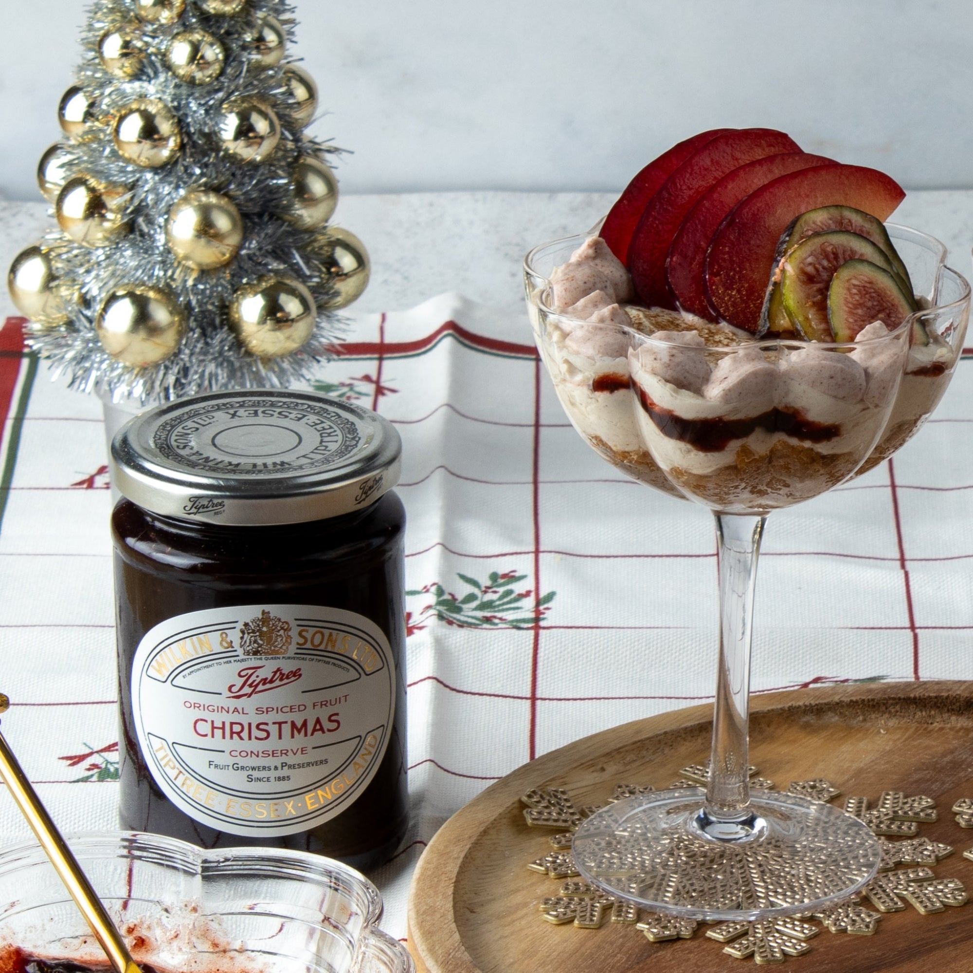 A glass filled with a layered pastry and sliced red fruit dessert, styled on a wooden tray beside a jar of Tiptree Christmas jam, a small silver Christmas tree, and a holiday tablecloth.