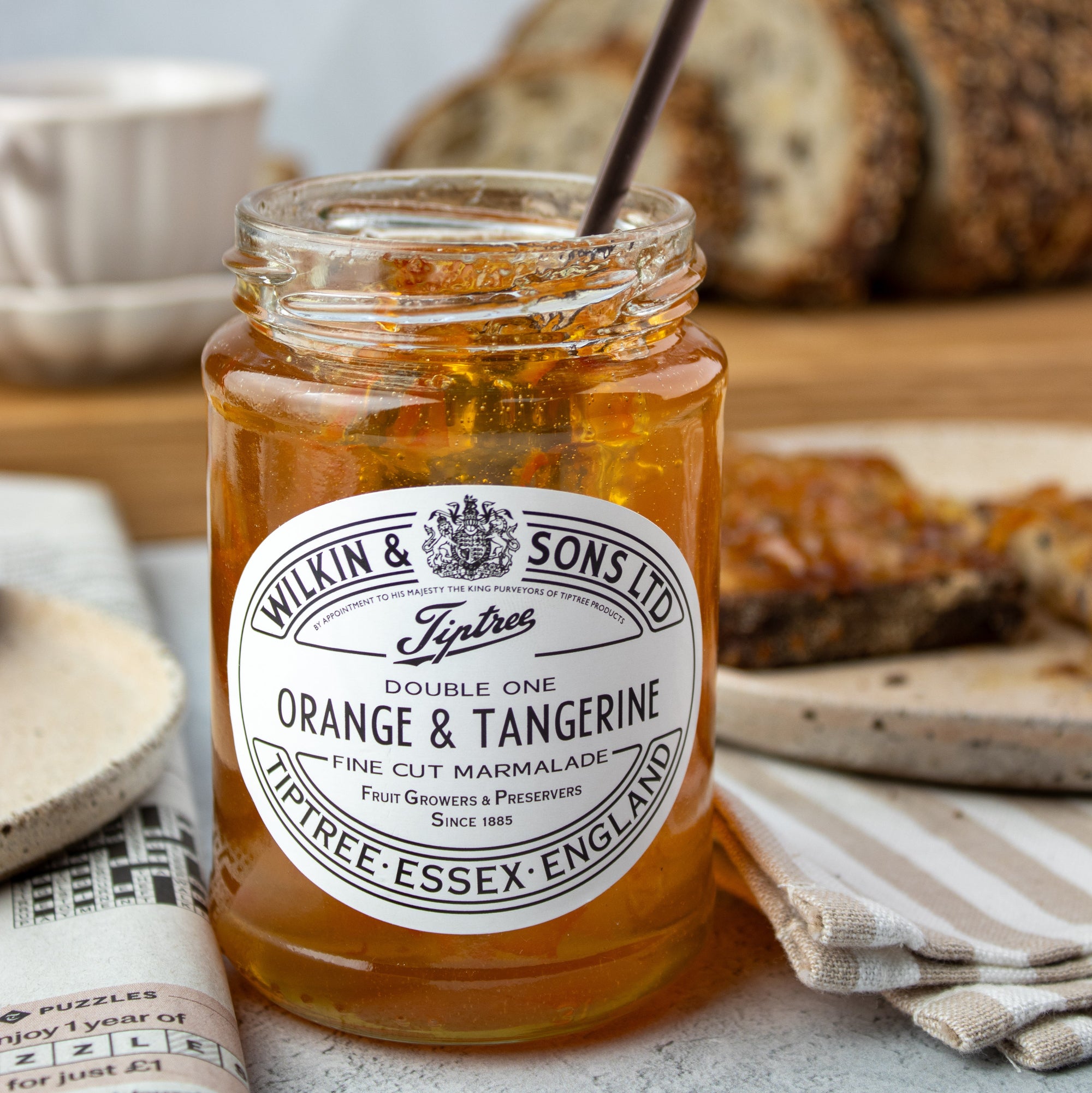 A glass jar of Tiptree Orange & Tangerine marmalade sits on a tabletop with sliced bread in the background. The amber-orange preserve is visible through the clear jar, with a dark spoon resting inside and a large white brand label on the front.