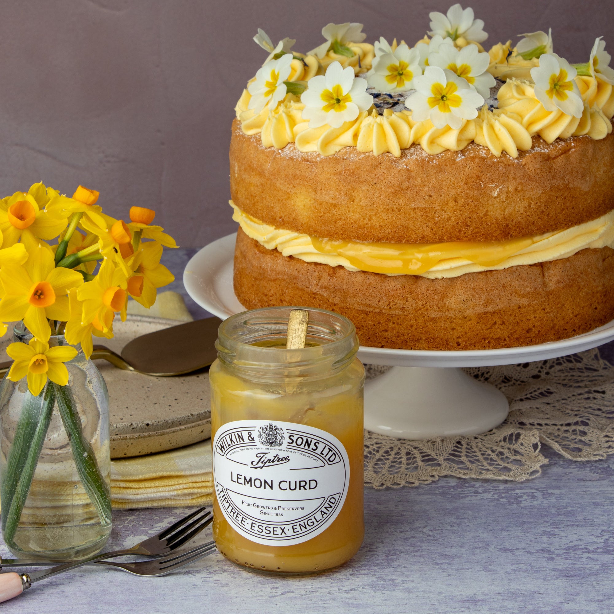 A two-tier sponge cake with yellow cream and white daffodils sits on a white cake stand, alongside a jar of Lemon Curd by Lemon & Sons and yellow daffodils in a glass vase.