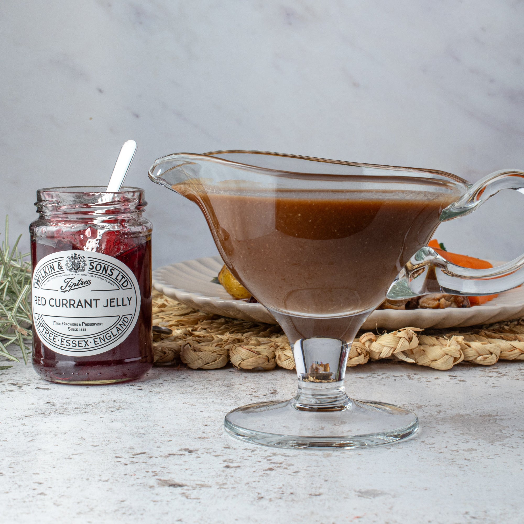 A clear glass gravy boat filled with gravy sits beside a glass jar of Tiptree Red Currant Jelly on a light stone surface, with woven placemats and a plate in the background.