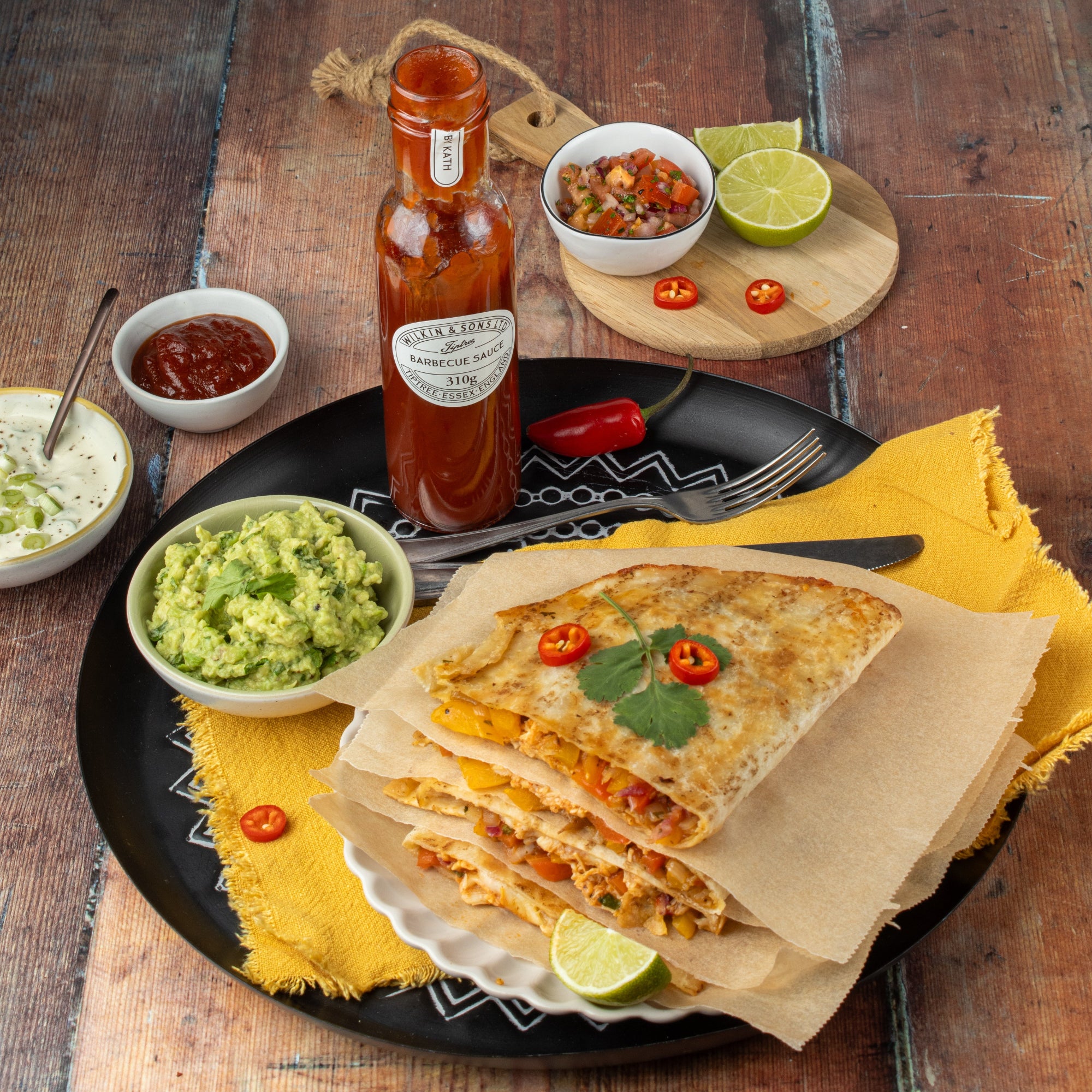 Quesadilla on a black plate with guacamole, salsa, and Tiptree barbecue sauce on a wooden table.