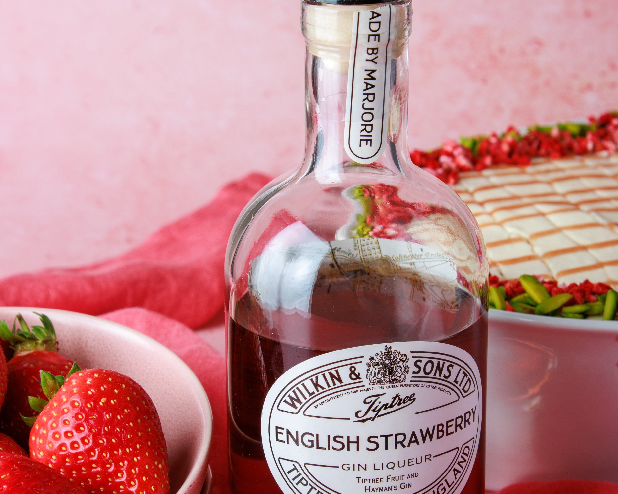 Bottle of Wilkin & Sons English Strawberry Gin Liqueur with strawberries and cake on a pink background