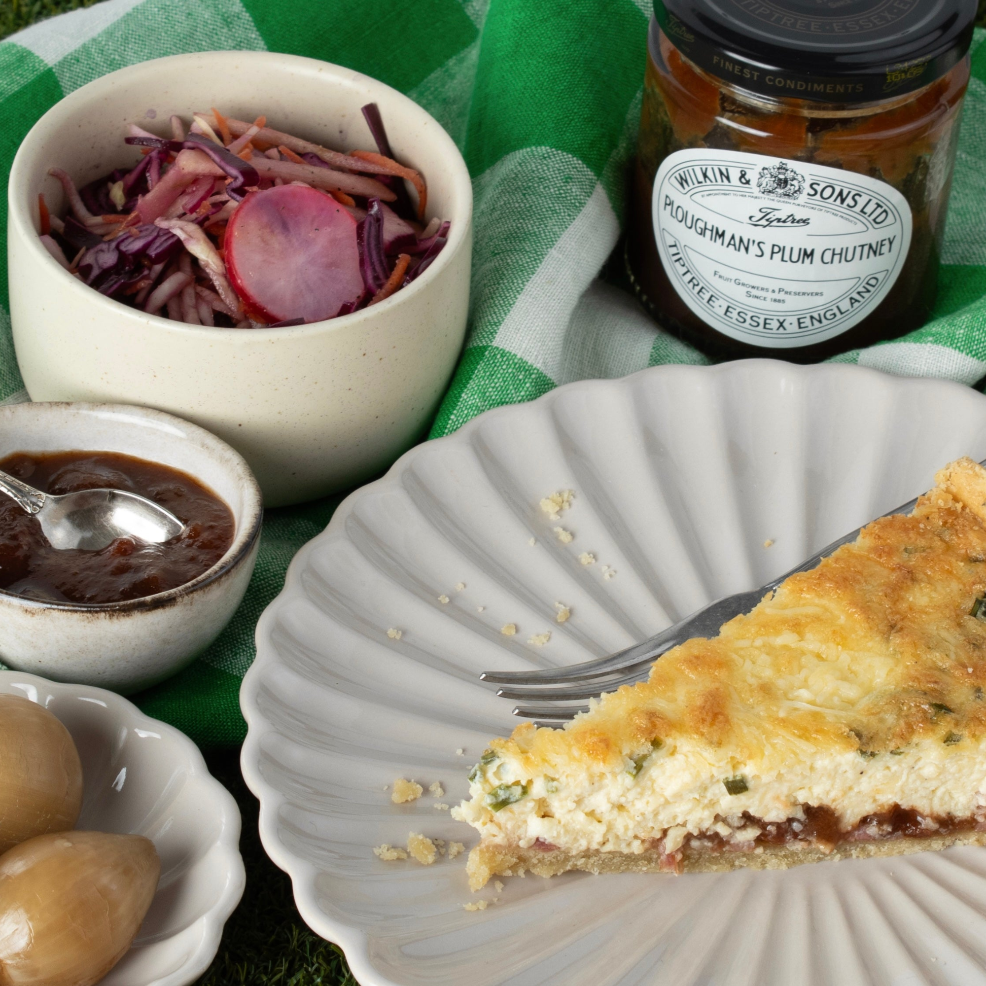 Slice of quiche on a plate with olives, a jar of plum chutney, and a bowl of pickled beets on a green checkered cloth.