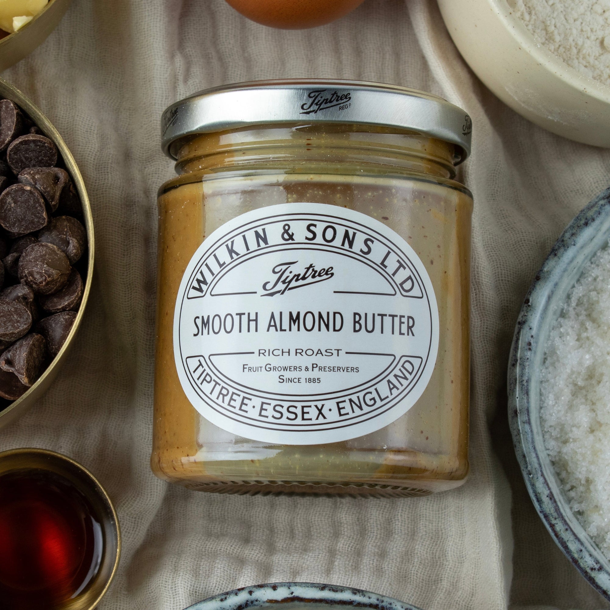 Jar of Wilkin & Sons Smooth Almond Butter surrounded by ingredients on a textured surface