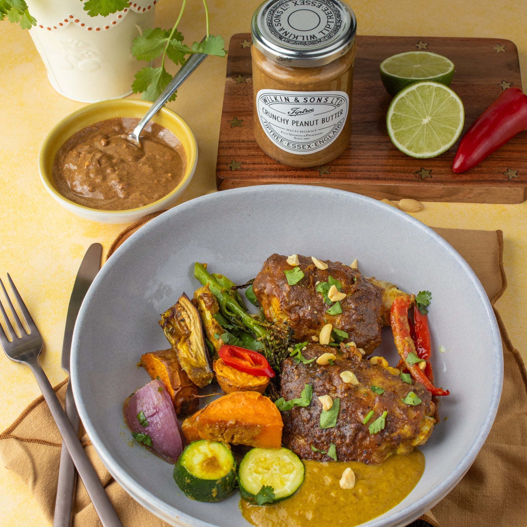 A plated dish of roasted chicken with vegetables in a creamy peanut sauce, served in a light blue bowl on a table with a jar of Wilkin & Sons Crunchy Peanut Butter, a small bowl of sauce, and garnish.
