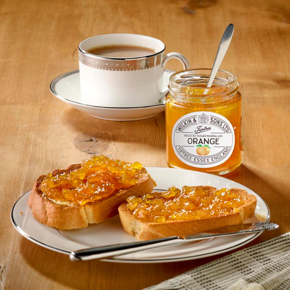 Jar of orange marmalade with a plate of toast spread with marmalade, a cup of tea, and a spoon on a wooden table.