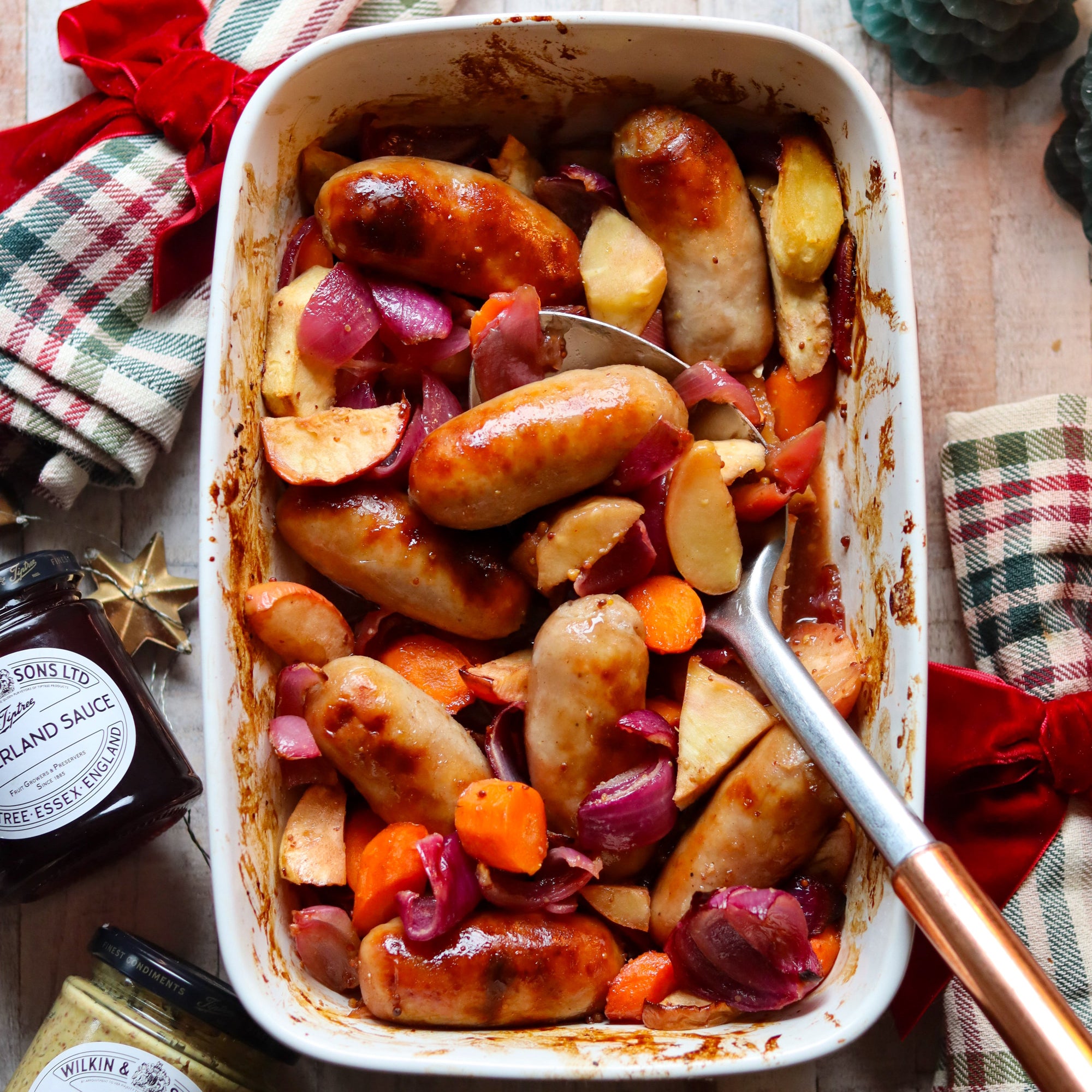 Baked sausages with vegetables in a white dish on a wooden table.