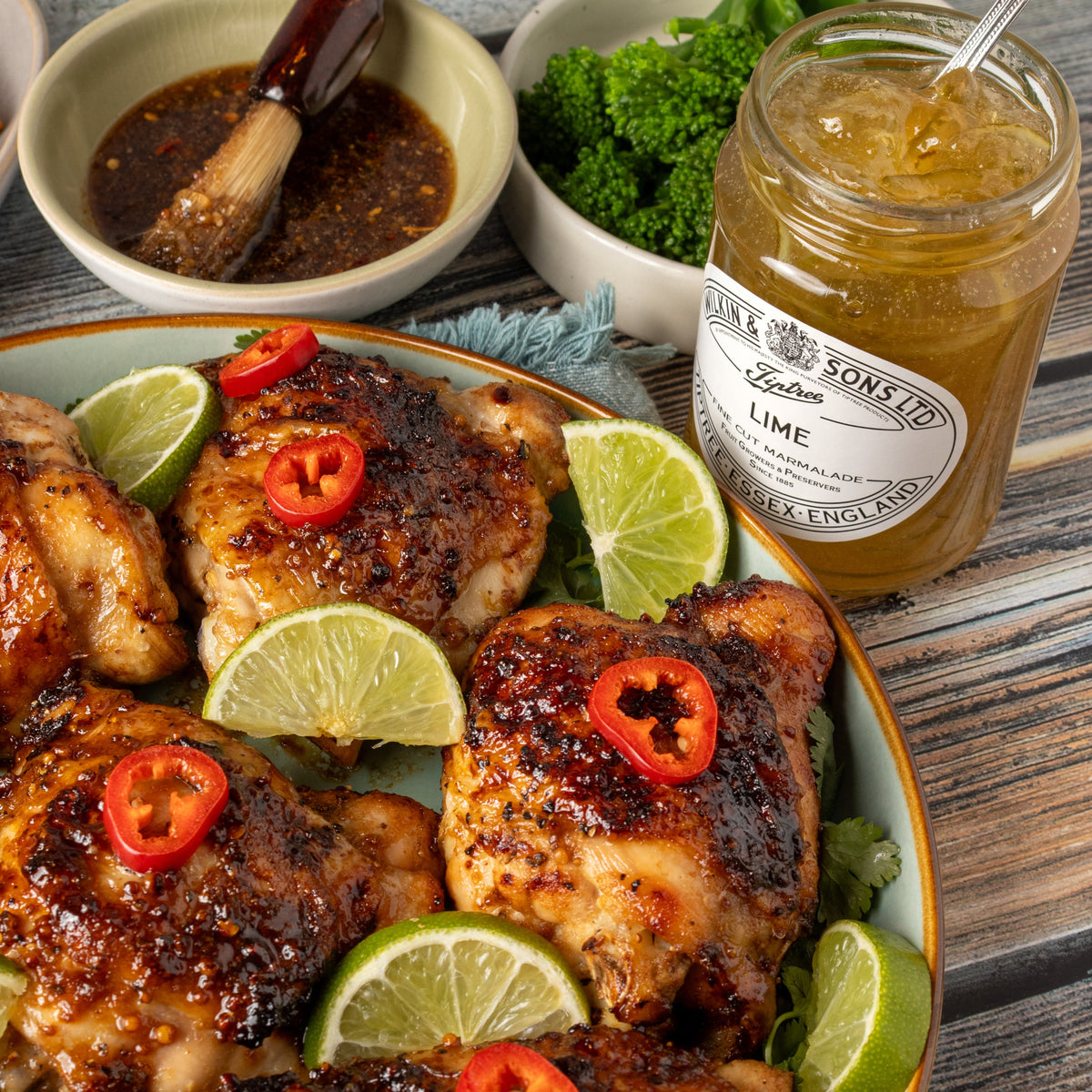 Grilled chicken pieces on a plate with lime slices and red chili slices, styled with bowls of dipping sauce and broccoli, next to a jar of Tiptree Lime Marmalade.