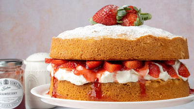 Image of a lavish Victoria Sponge filled with Strawberries and Cream next to a jar of Wilkin & Sons Tiptree Strawberry Jam