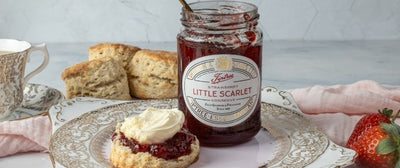 A scone with Jam and Clotted Cream in the correct order next to a jar of Tiptree Little Scarlet Strawberry Conserve
