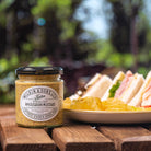 Jar of Wilkin & Sons English Wholegrain Mustard on a wooden table with a plate of sandwiches and fruit in the background.