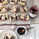 Black Currant cheesecake bars arranged on parchment paper, with an open jar and a small bowl of dark Black Currant jam, and a plated slice on a white tabletop.