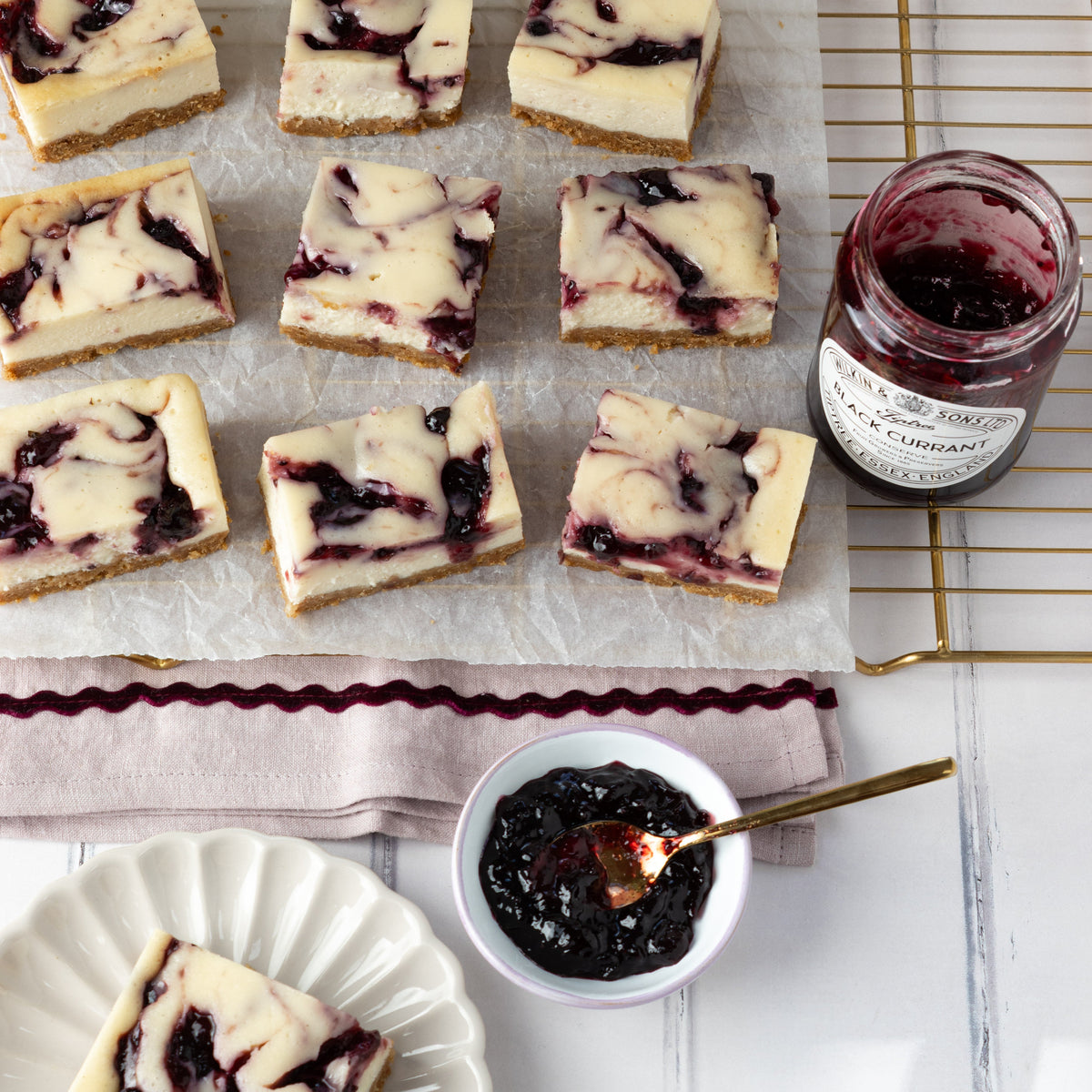 Black Currant cheesecake bars arranged on parchment paper, with an open jar and a small bowl of dark Black Currant jam, and a plated slice on a white tabletop.