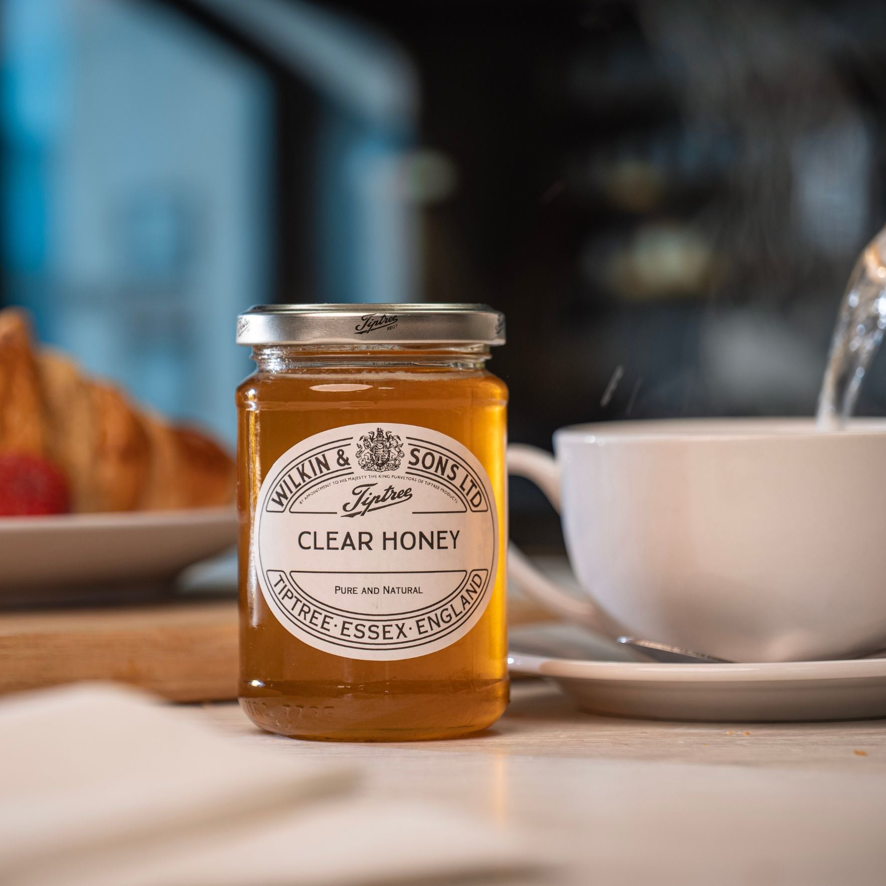Jar of Wilkin & Sons Clear Honey on a table with a cup and croissant in the background