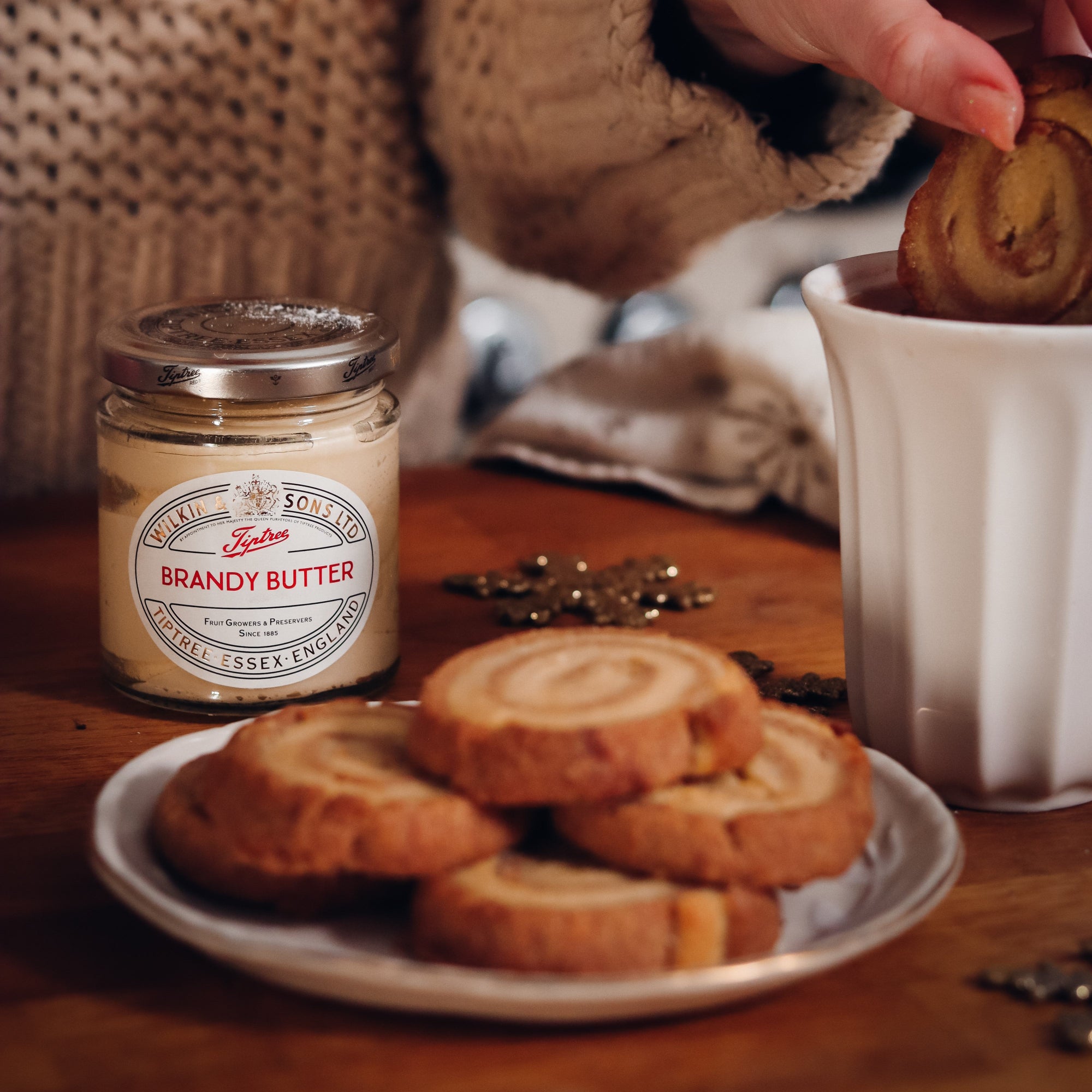 A jar of Brandy Butter with biscuits on a wooden table, with a hand dipping a biscuit into a white mug.