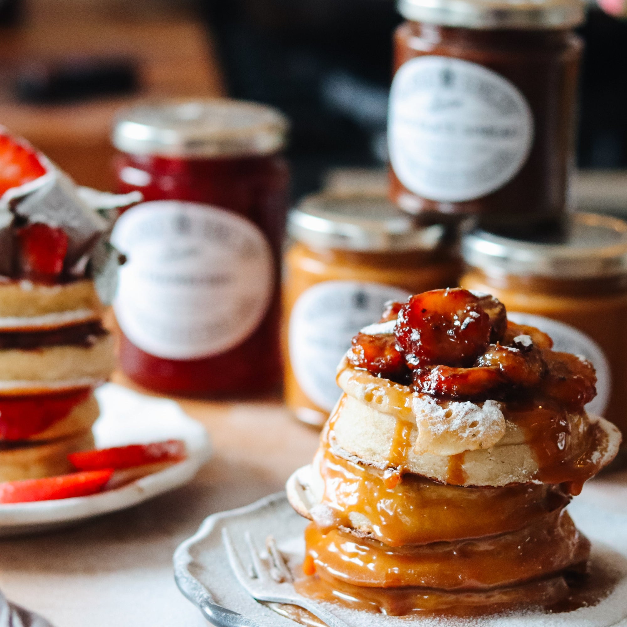 A close-up food scene showing a tall stack of pancakes topped with glossy caramel sauce and fruit, with several jars of jam or preserve in the background on a tabletop.