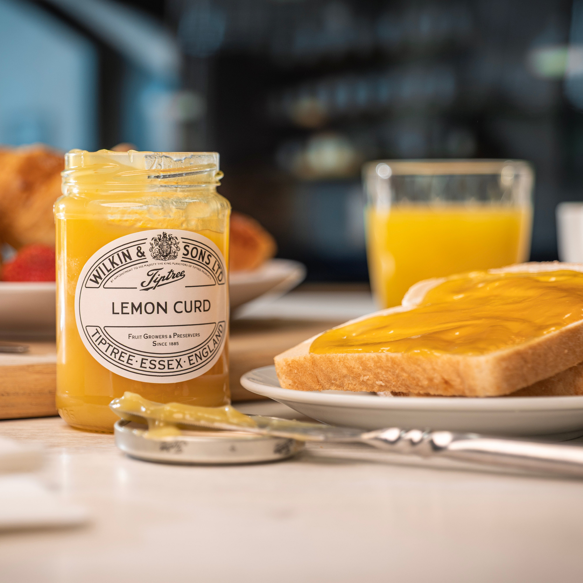 Jar of lemon curd with a slice of toast and orange juice on a table.