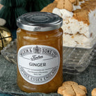 A jar of Wilkin & Sons Ltd Ginger Conserve on a dark textured cloth, with gingerbread cookies and a decorated Christmas cake in the background.