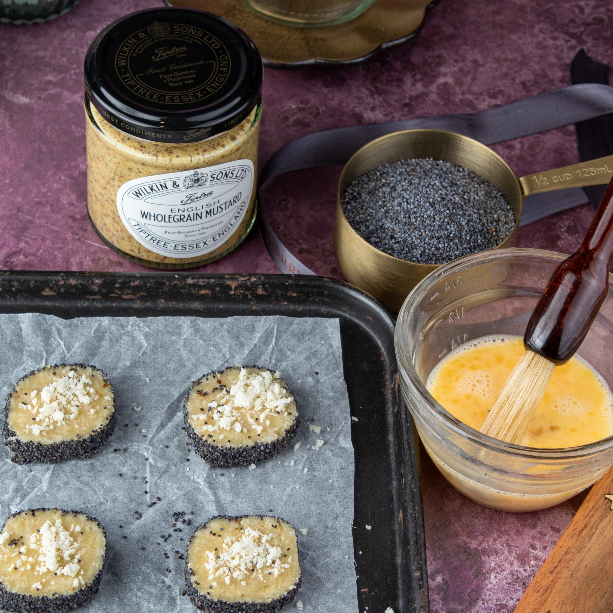 A styled food preparation scene showing several round cheese nibbles on a baking tray, a glass bowl of beaten egg with a whisk, a small cup of poppy seeds, and a jar labeled Wilkin & Sons Wholegrain Mustard on a purple tabletop.