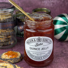 A jar of Wilkin & Sons Ltd Quince Jelly on a textured purple surface, surrounded by poppy seed cookies, a gold spoon, and other jars in the background.