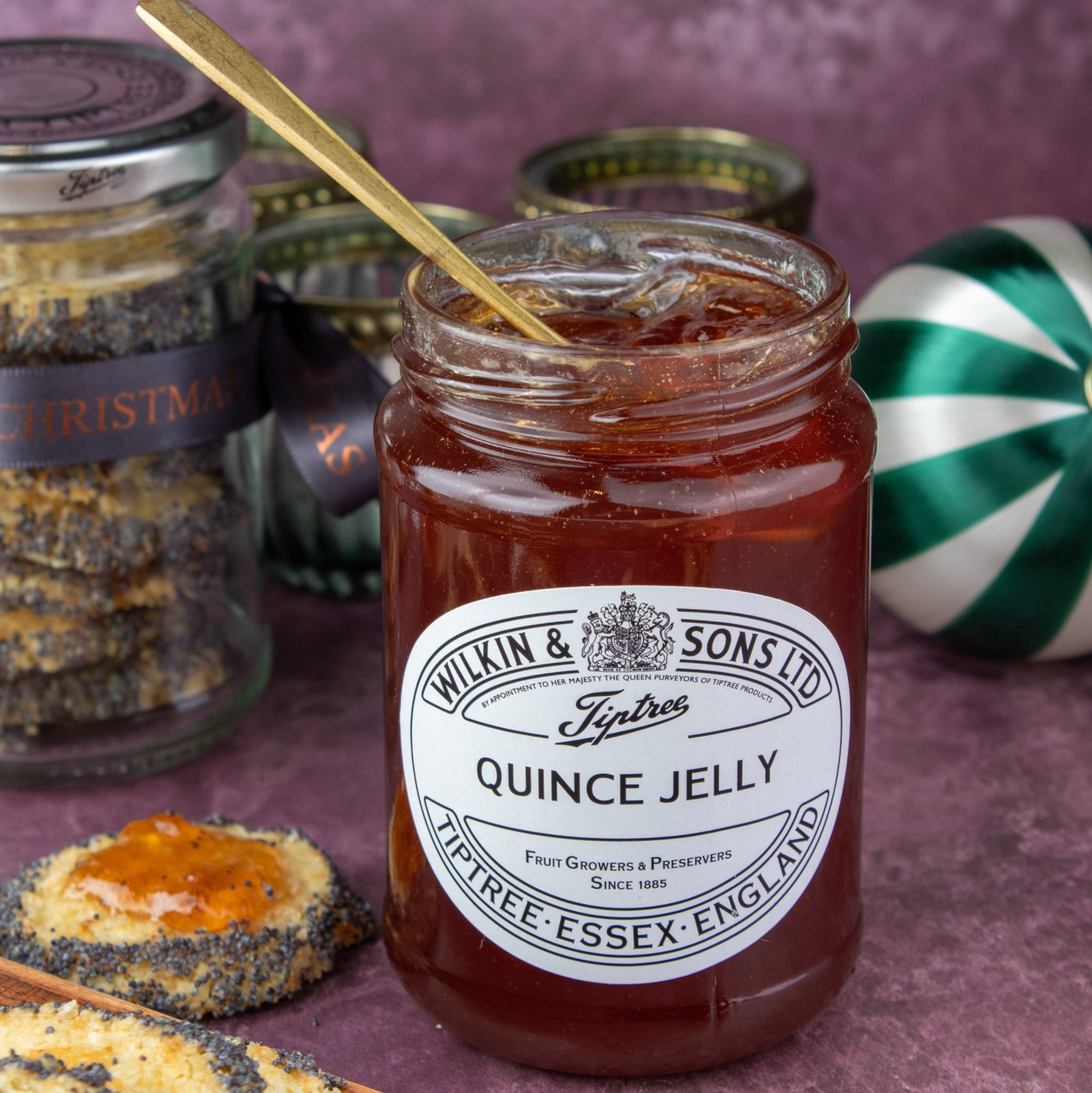A jar of Wilkin & Sons Ltd Quince Jelly on a textured purple surface, surrounded by poppy seed cookies, a gold spoon, and other jars in the background.