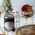 A glass filled with a layered pastry and sliced red fruit dessert, styled on a wooden tray beside a jar of Tiptree Christmas jam, a small silver Christmas tree, and a holiday tablecloth.