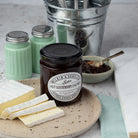A jar of Tiptree Hot Gooseberry Chutney displayed on a stone plate with cheese, salt shakers, a silver bucket with cutlery, and a small bowl of chutney spread on a light stone surface.