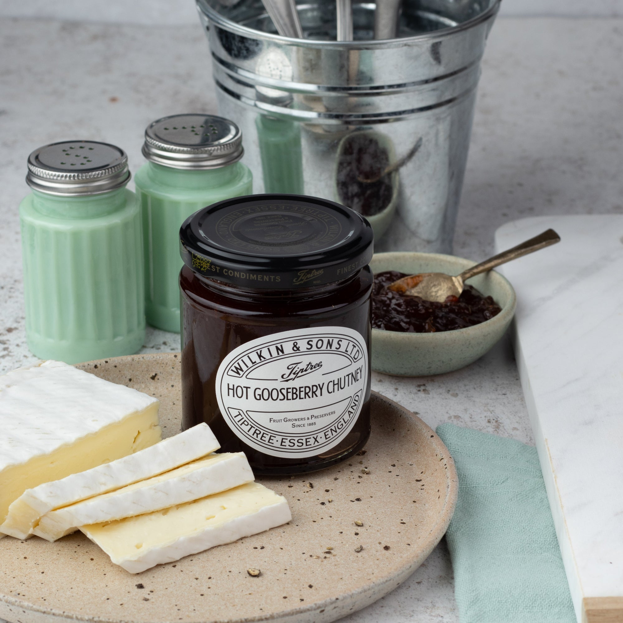 A jar of Tiptree Hot Gooseberry Chutney displayed on a stone plate with cheese, salt shakers, a silver bucket with cutlery, and a small bowl of chutney spread on a light stone surface.