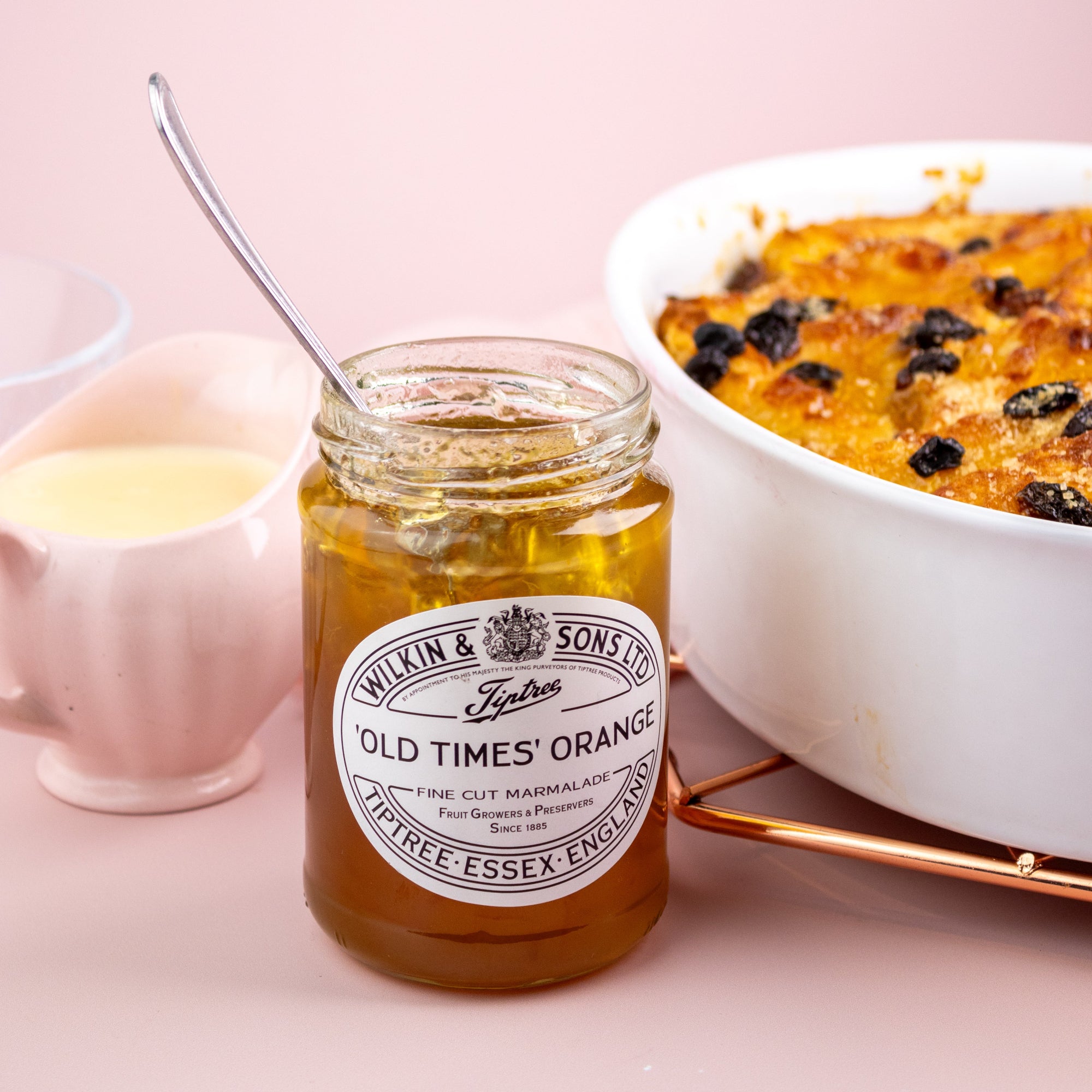 Jar of Wilkin & Sons 'Old Times' Orange Marmalade with a bowl of dessert and a glass on a pink background