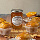 Jar of Wilkin & Sons Crystal Orange marmalade with muffins on a wooden surface