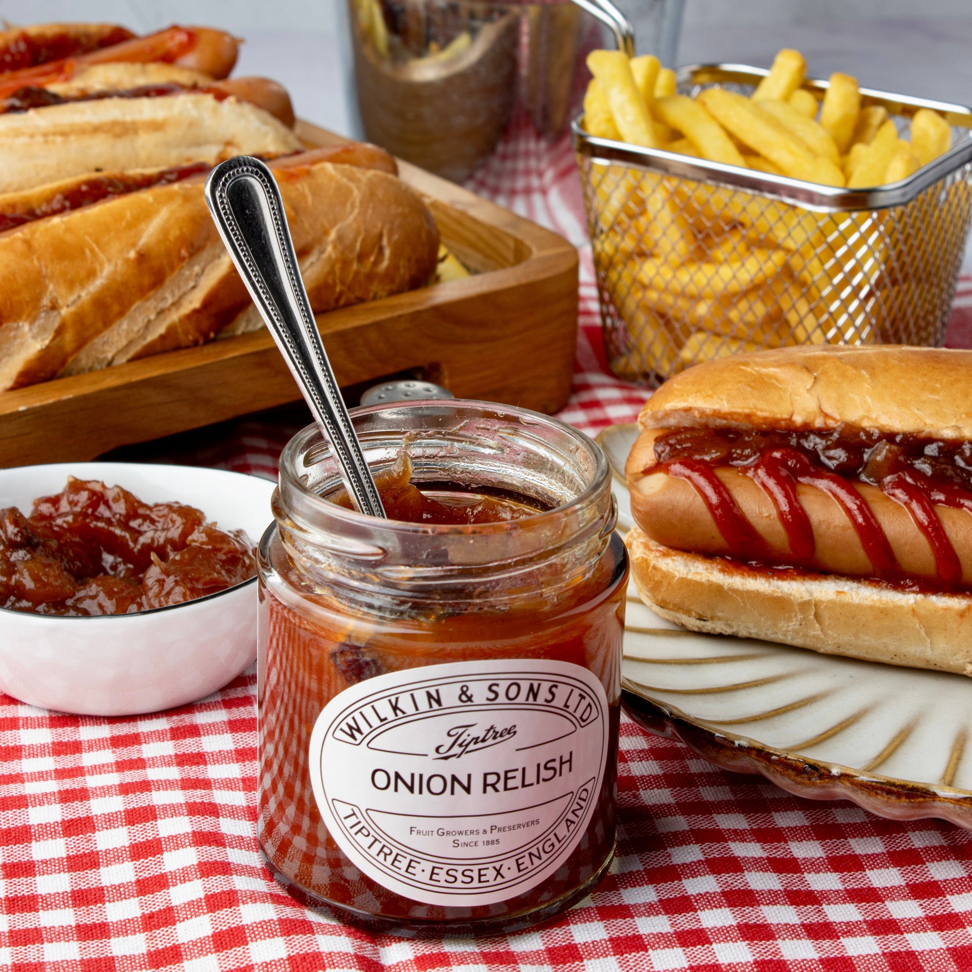 Jar of onion relish with a spoon, hot dogs, and fries on a checkered tablecloth.