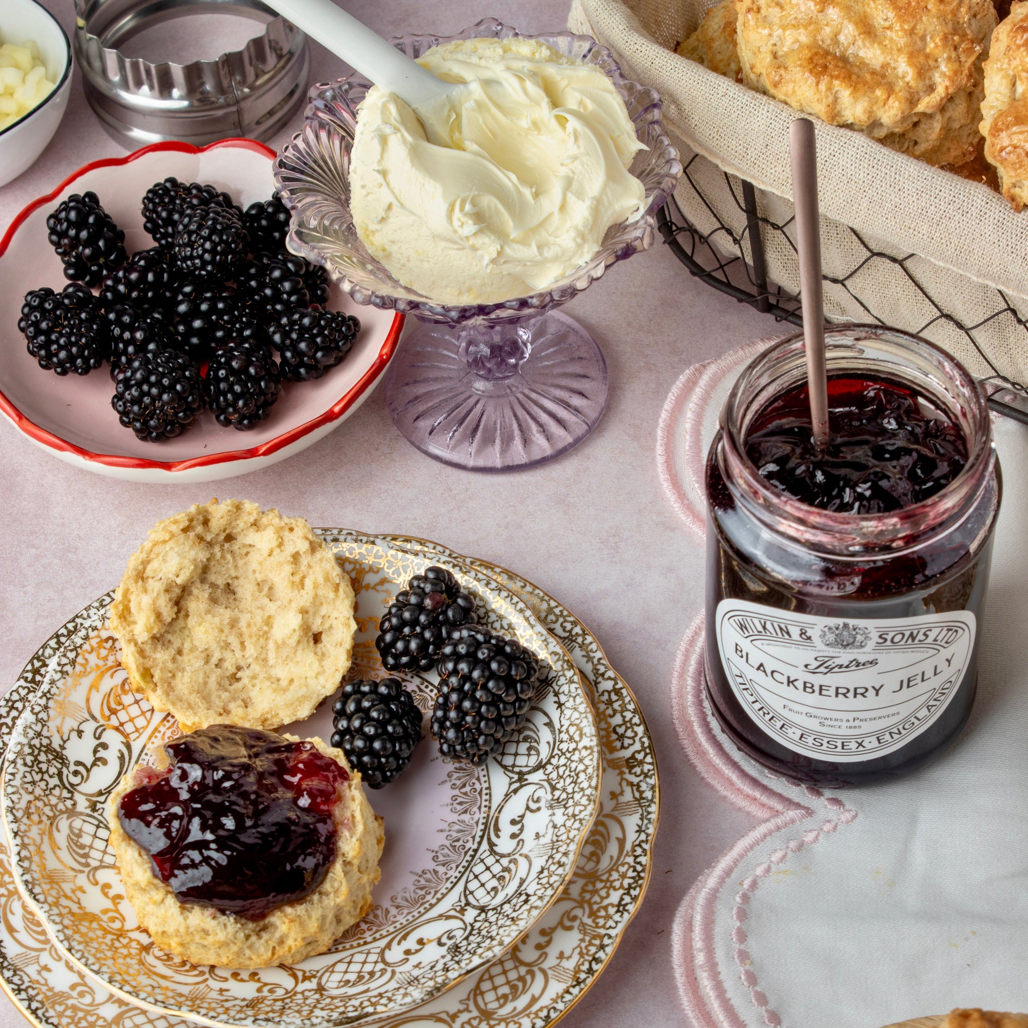 A styled tabletop scene showing a jar of Tiptree Blackberry Jelly alongside blackberries, scones, and a bowl of whipped cream on a pale pink surface.