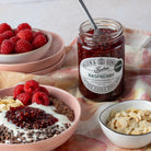 A glass jar of Wilkin & Sons Tipton Organic Raspberry Conserve styled on a floral cloth with bowls of raspberries, yogurt, chocolate, and sliced almonds.