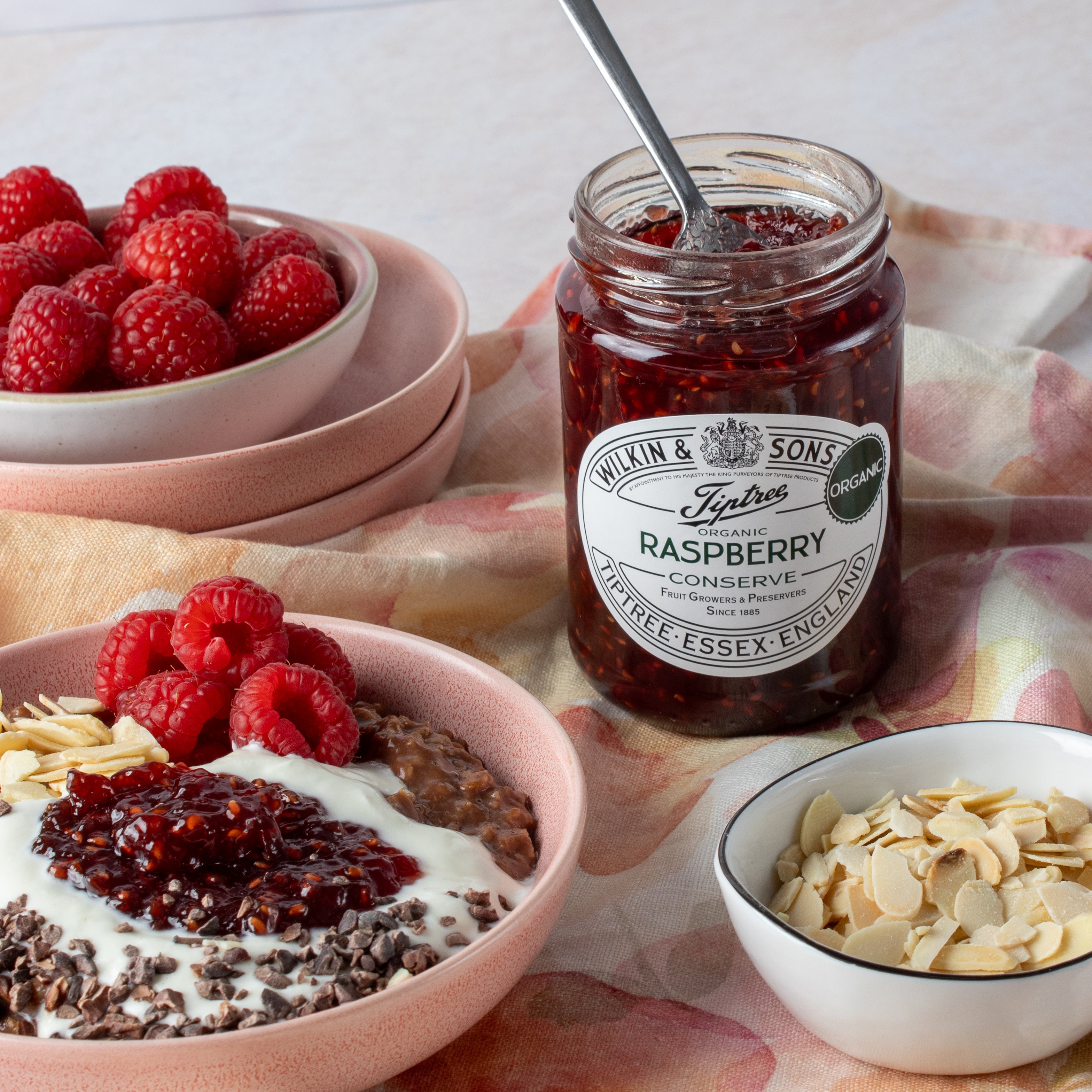 A glass jar of Wilkin & Sons Tipton Organic Raspberry Conserve styled on a floral cloth with bowls of raspberries, yogurt, chocolate, and sliced almonds.