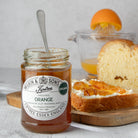 A jar of Wilkin & Sons organic orange marmalade sits on a wooden board with a spoon, alongside slices of bread spread with orange marmalade and an orange juicer in the background.