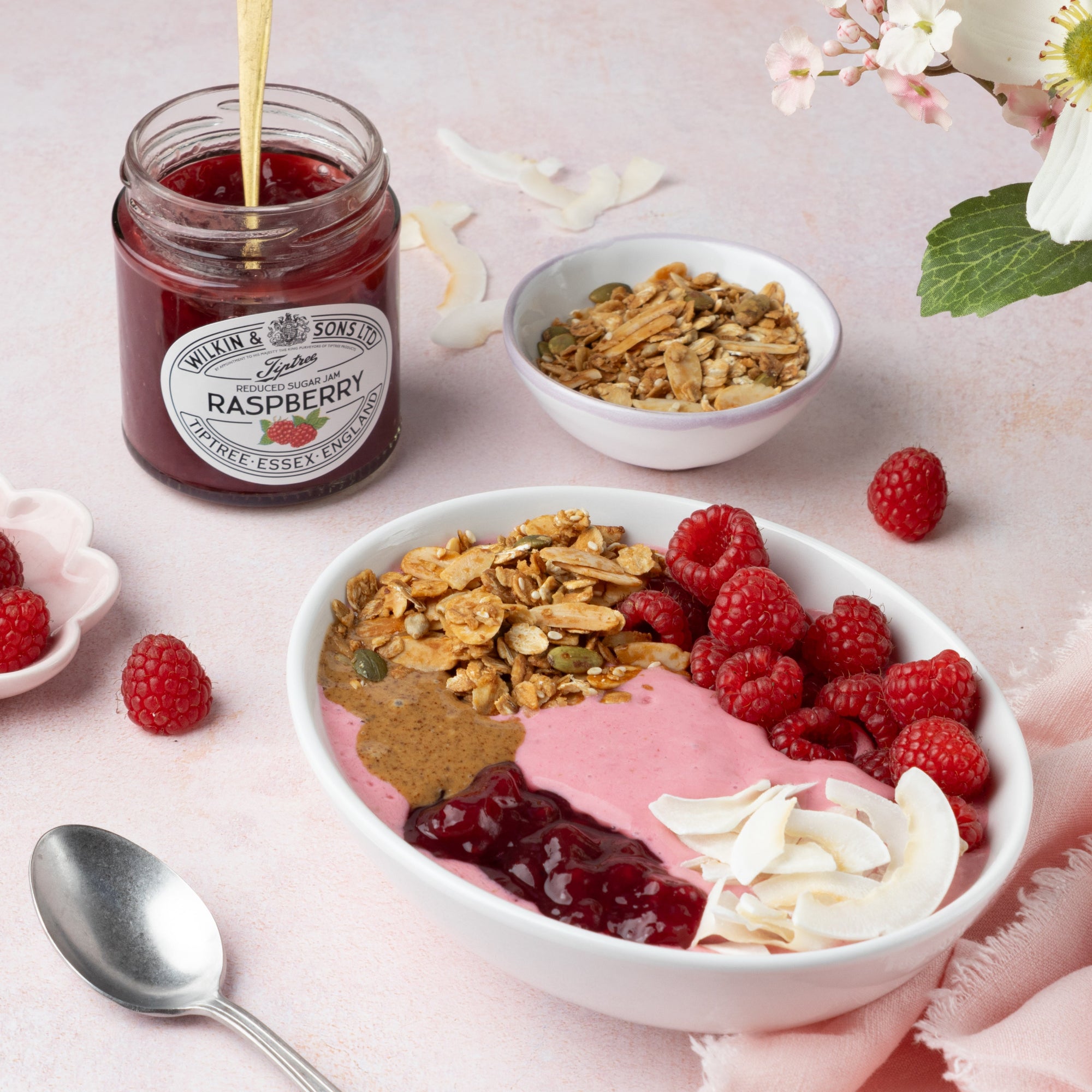 Bowl of yogurt with granola, raspberries, and a jar of raspberry jam on a light pink background.