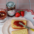 A jar of East Anglian Strawberry Jam sits on a pink tiled surface next to two whole strawberries and a plate with a slice of cake spread with jam and a knife.