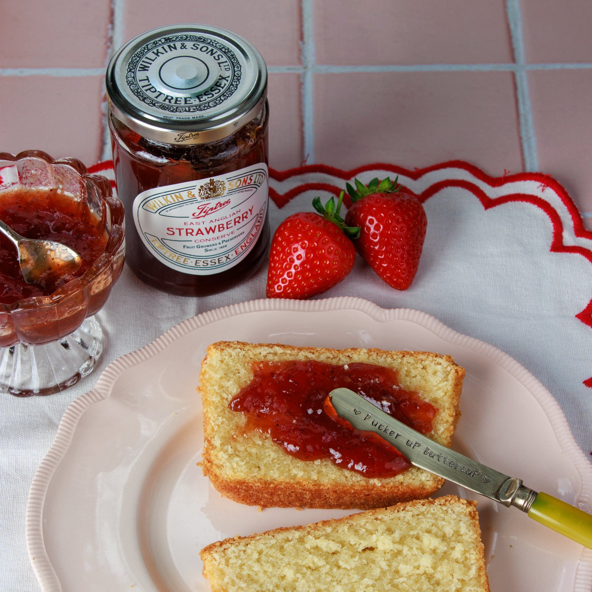A jar of East Anglian Strawberry Jam sits on a pink tiled surface next to two whole strawberries and a plate with a slice of cake spread with jam and a knife.