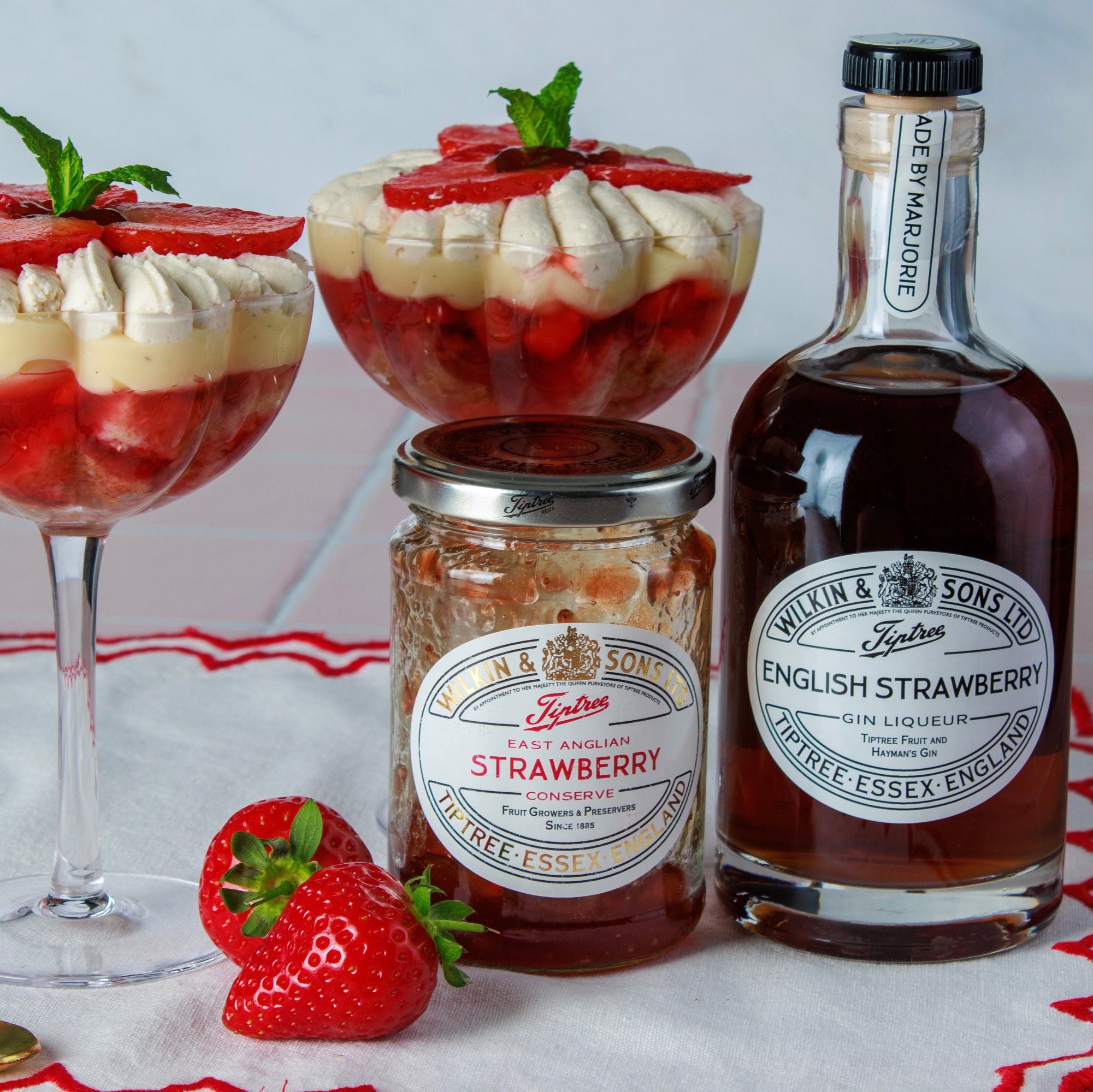 Two strawberry desserts in glasses with a jar and bottle of Pickles & Son's English Strawberry Preserves on a red and white checkered tablecloth.