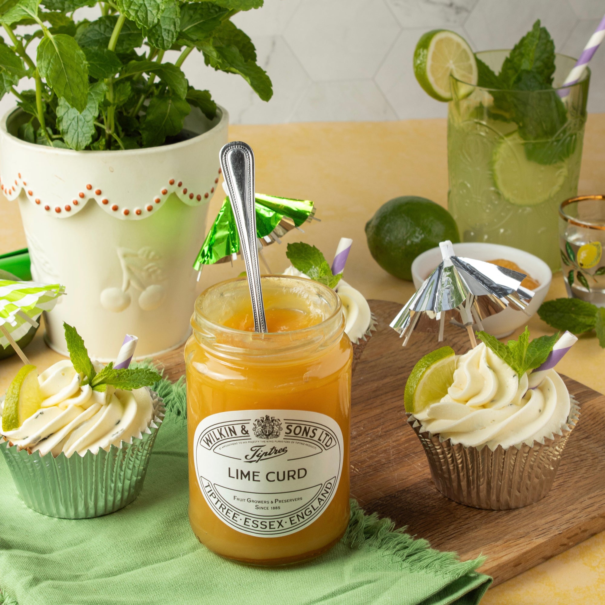 Jar of lime curd with a spoon, surrounded by cupcakes and a glass of limeade on a wooden board.