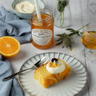Slice of cake with Greek yoghurt on a plate, jar of orange with malt whiskey marmalade, half an orange, and a glass of drink on a table.
