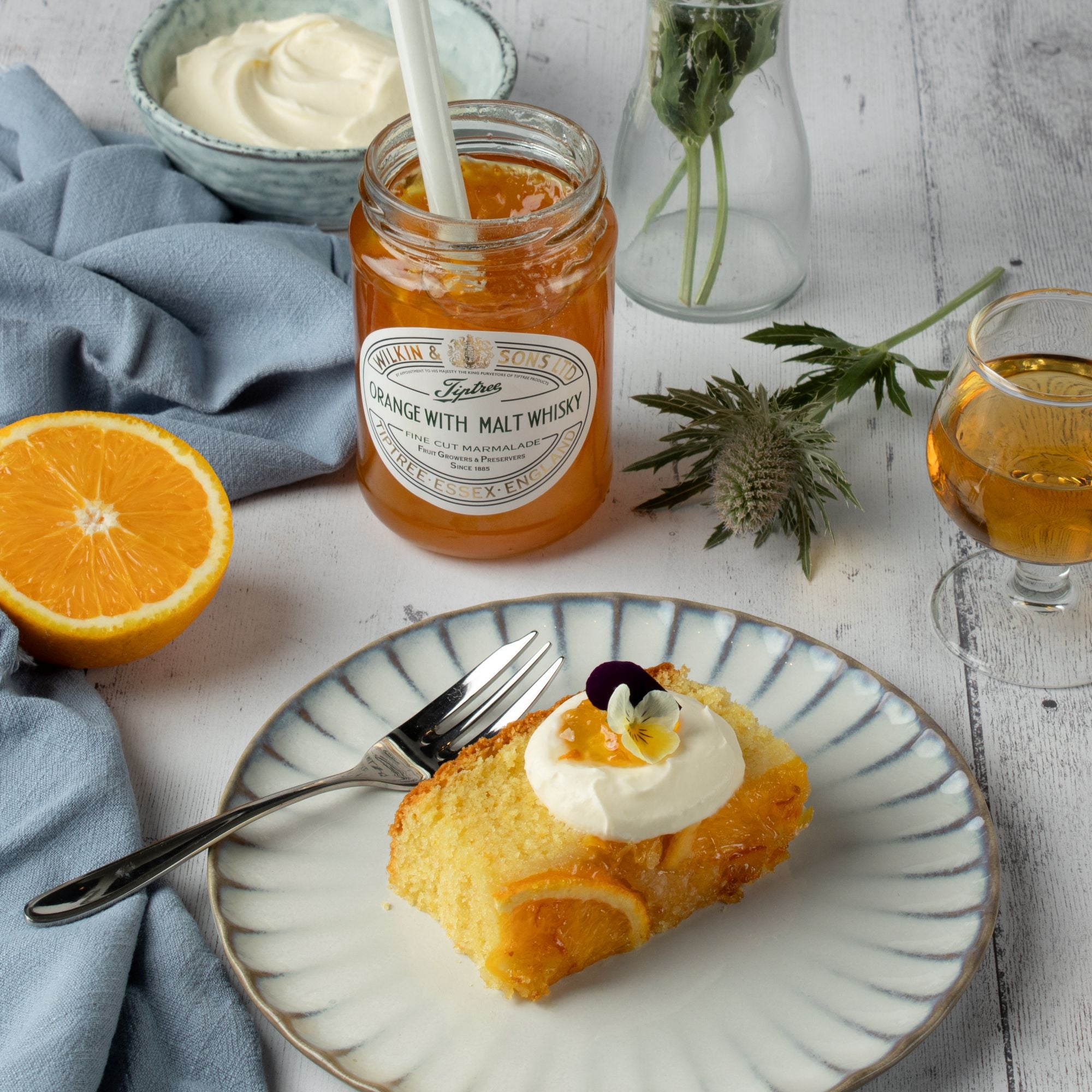 Slice of cake with Greek yoghurt on a plate, jar of orange with malt whiskey marmalade, half an orange, and a glass of drink on a table.