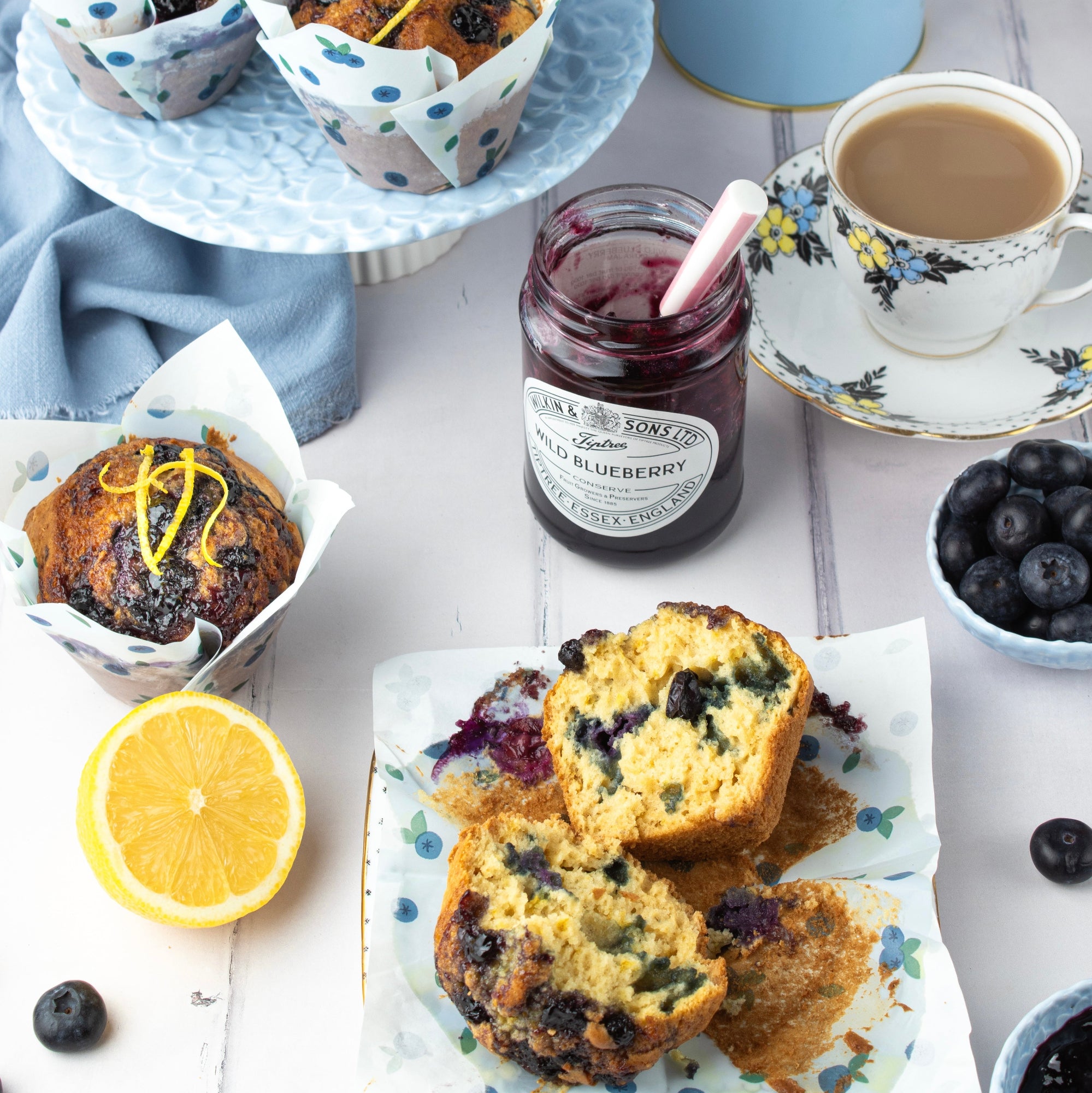 Blueberry muffins and a blueberry jam jar arranged on a white table with a cup of tea, blueberries, and a lemon half.