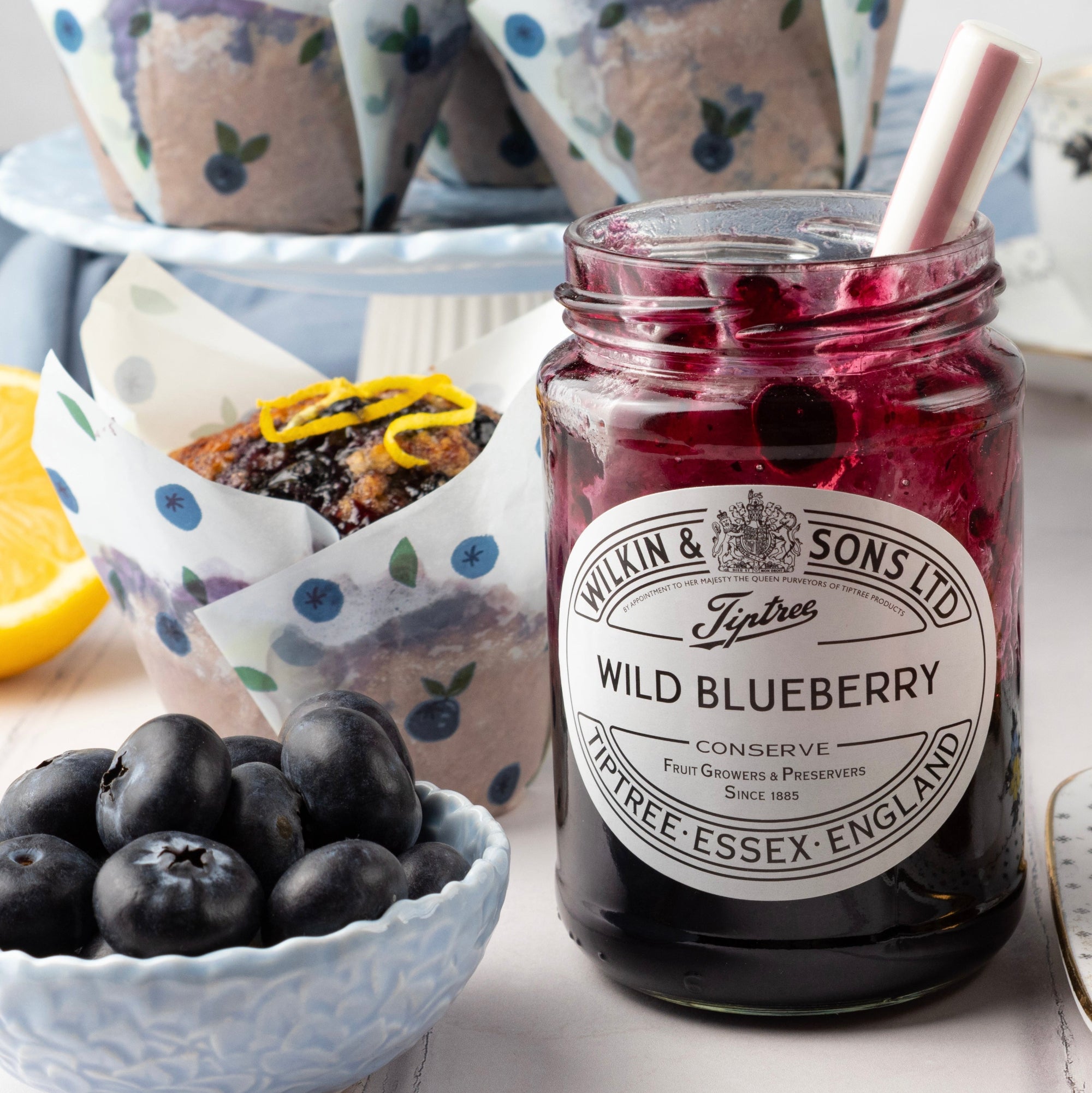 A jar of Wilkin & Sons Tiptree Wild Blueberry Conserve in the foreground, with blueberries in a bowl, muffins in paper liners on a stand, and a lemon slice on a tabletop.