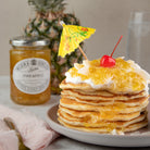 A tall stack of golden pancakes topped with whipped cream, bright yellow pineapple jam, a red cherry, and a small yellow cocktail umbrella, served on a plate with a jar of Wilkin & Sons pineapple jam in the background.