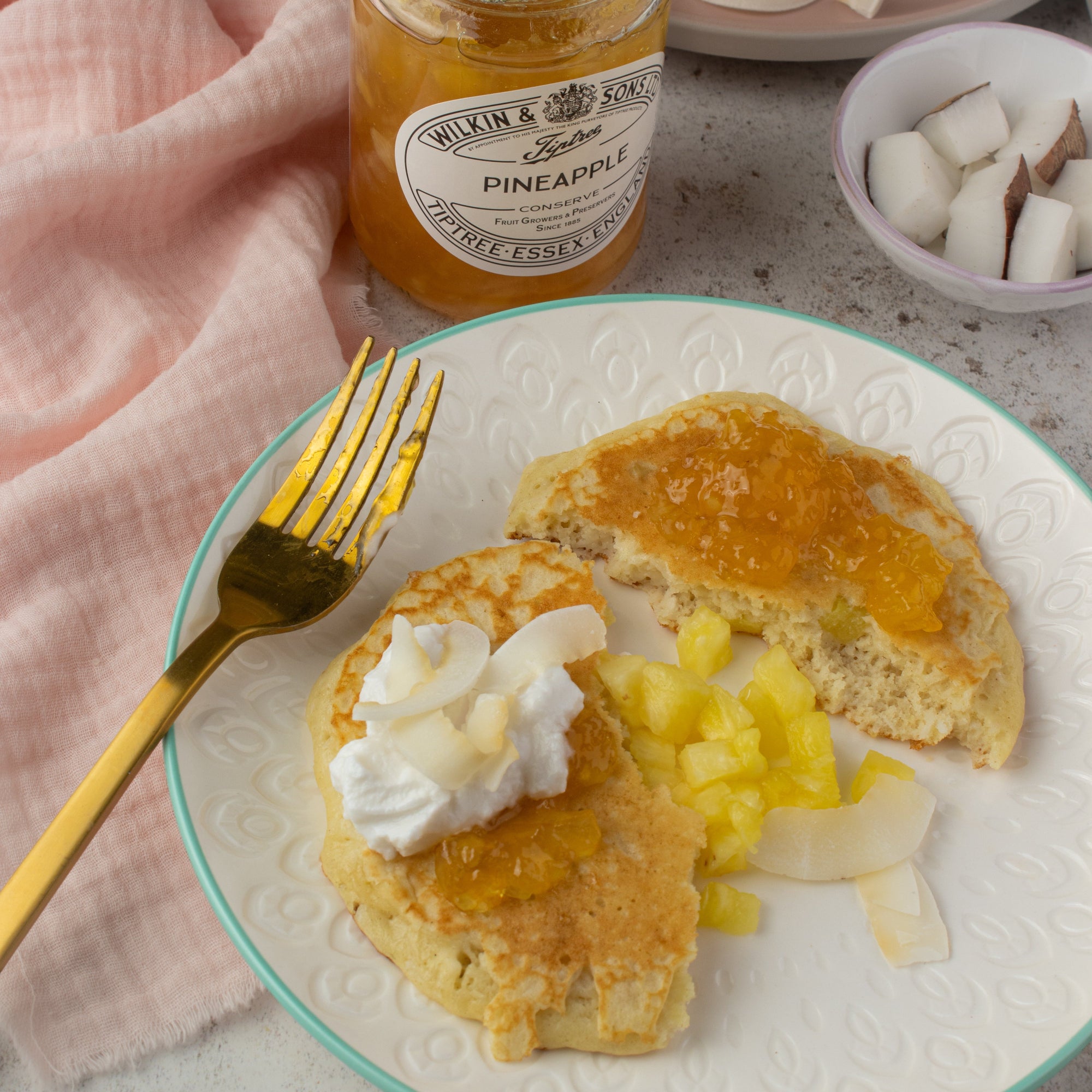 A styled breakfast scene showing a plate with two pancakes topped with pineapple jam, whipped cream, and pineapple chunks, alongside a gold fork. A jar of Tiptree Pineapple Jam is visible in the background along with a pink napkin and a small bowl of coconut.